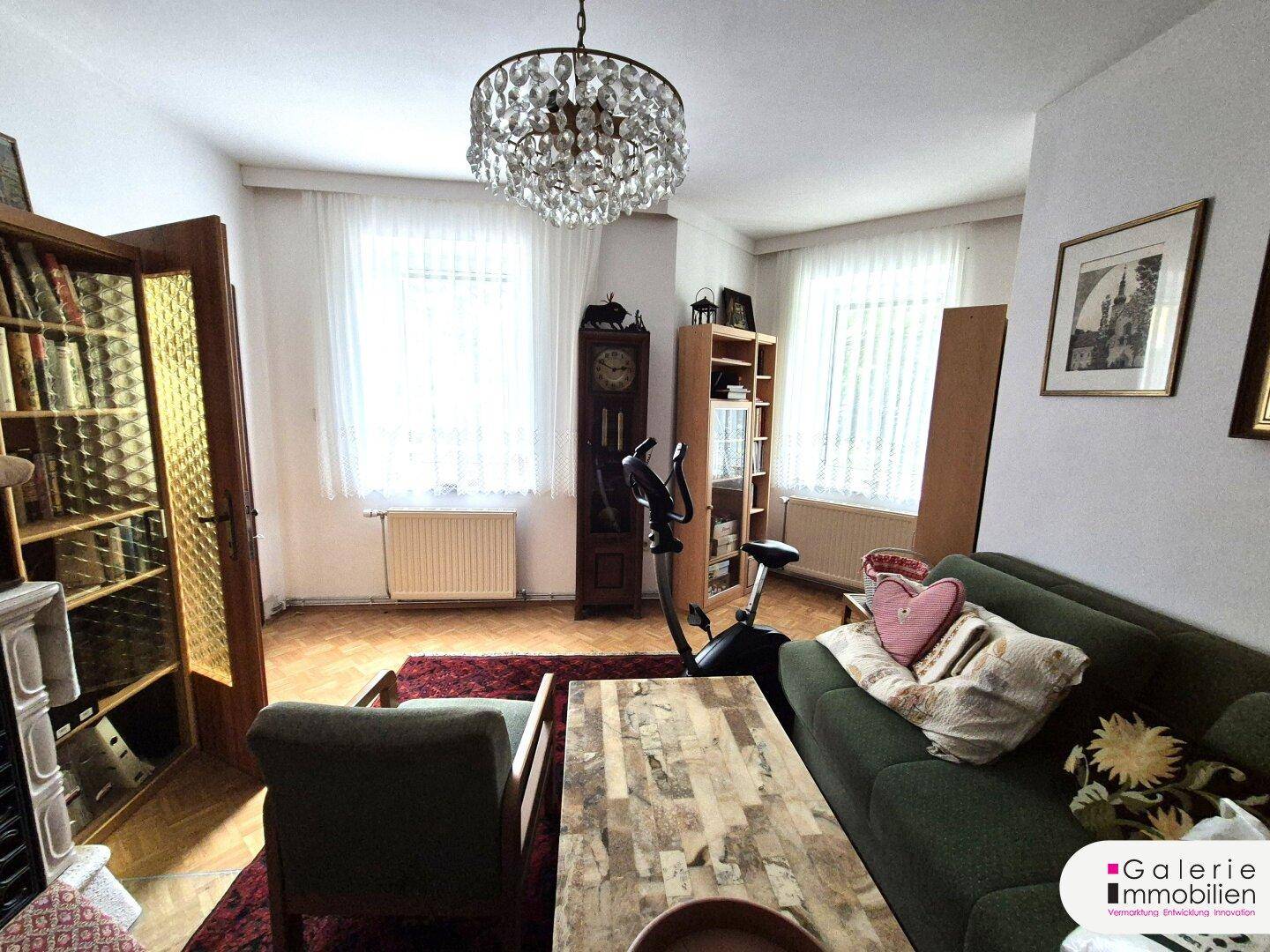 Helles Wohnzimmer mit Parkettboden, zwei Fenstern und einem Kronleuchter.