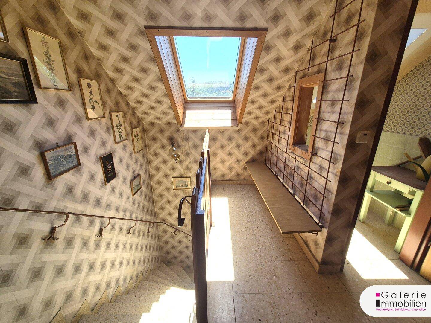Treppenhaus mit gemusterter Tapete, Holzgeländer und Dachfenster für natürliches Licht.