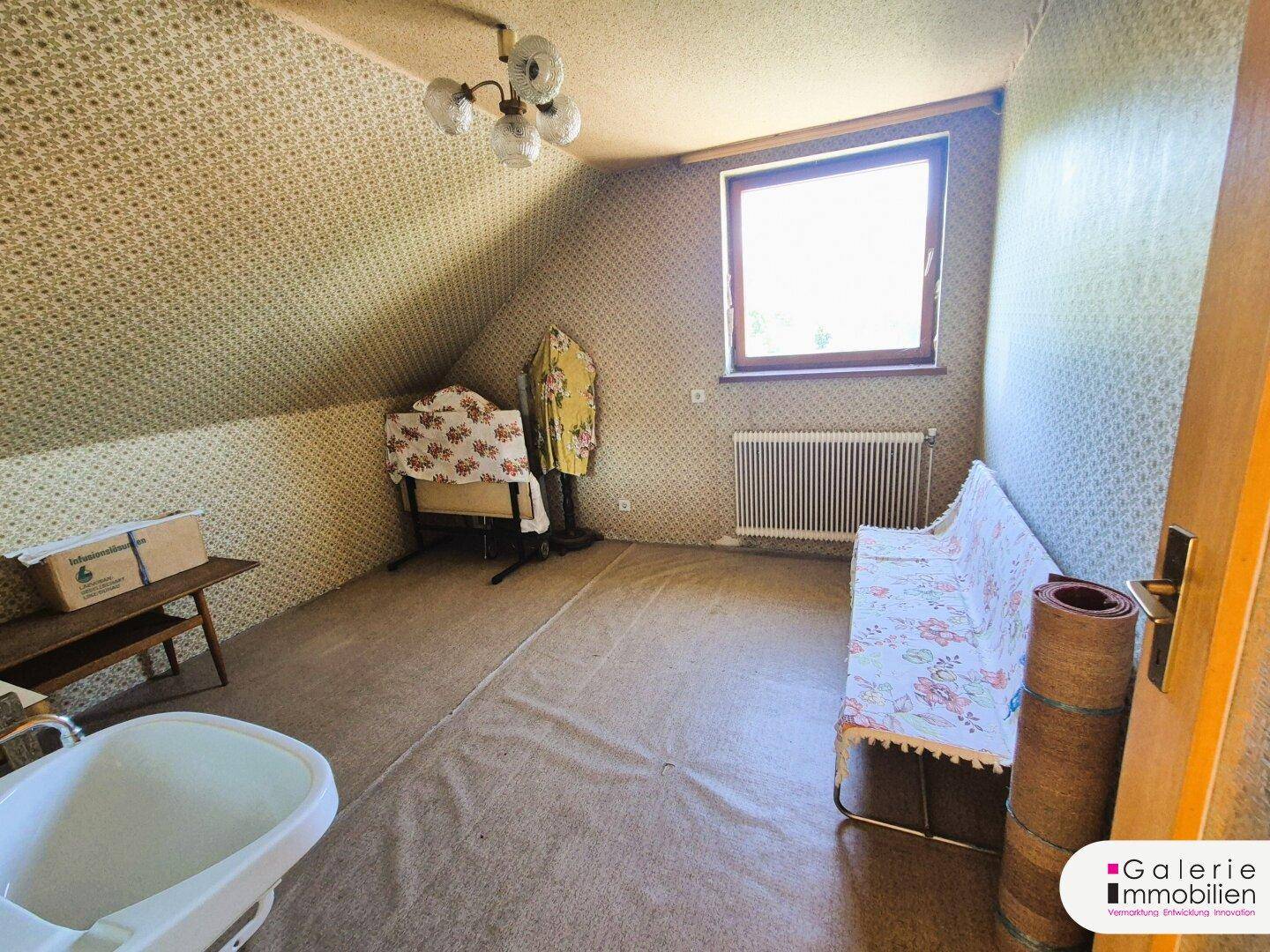 Dachzimmer mit schrägen Wänden, gemusterter Tapete und einem Fenster mit Blick ins Grüne.