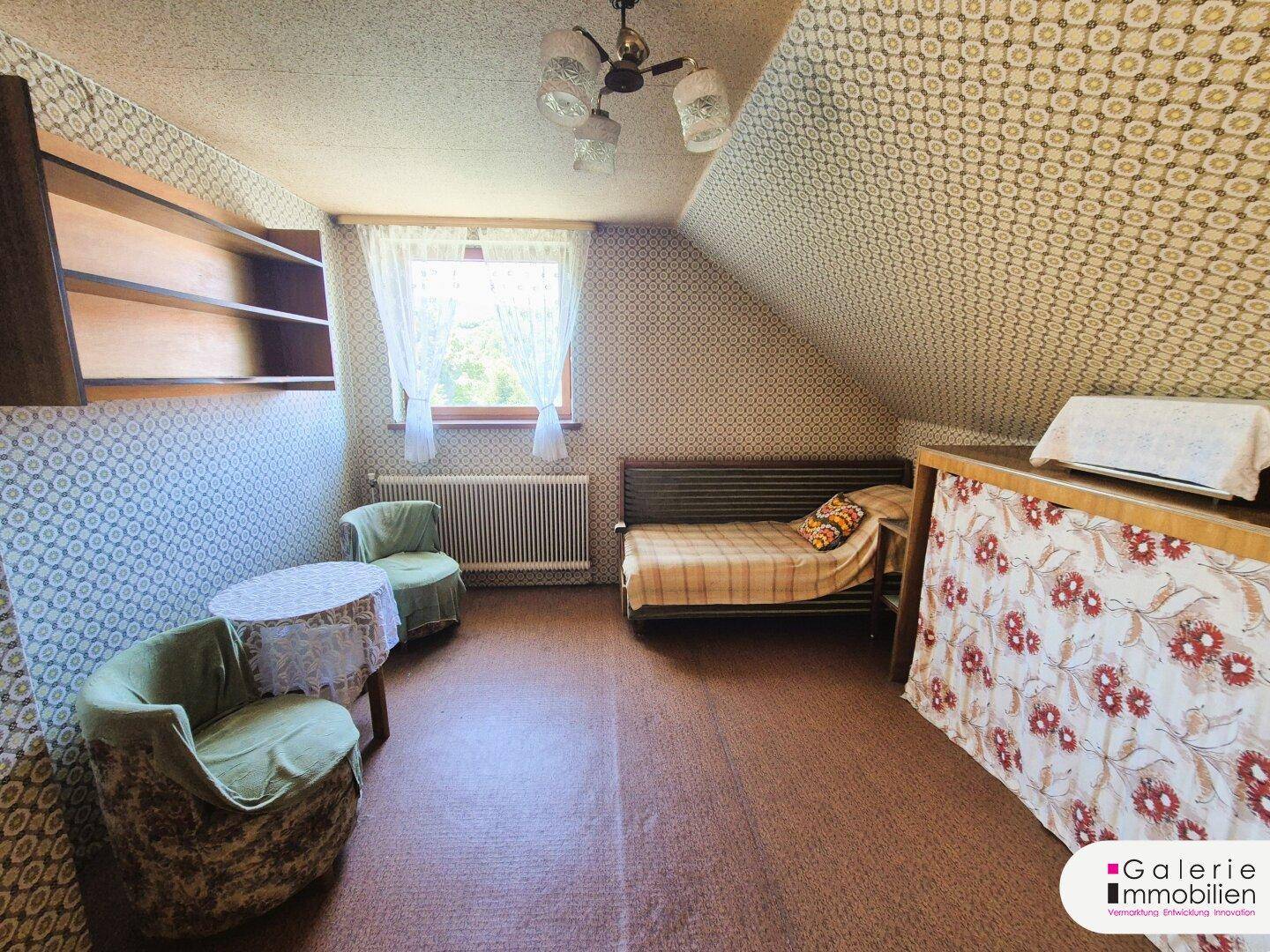 Weiteres Dachzimmer mit schrägen Wänden, Sitzgelegenheiten und Blick aus dem Fenster.