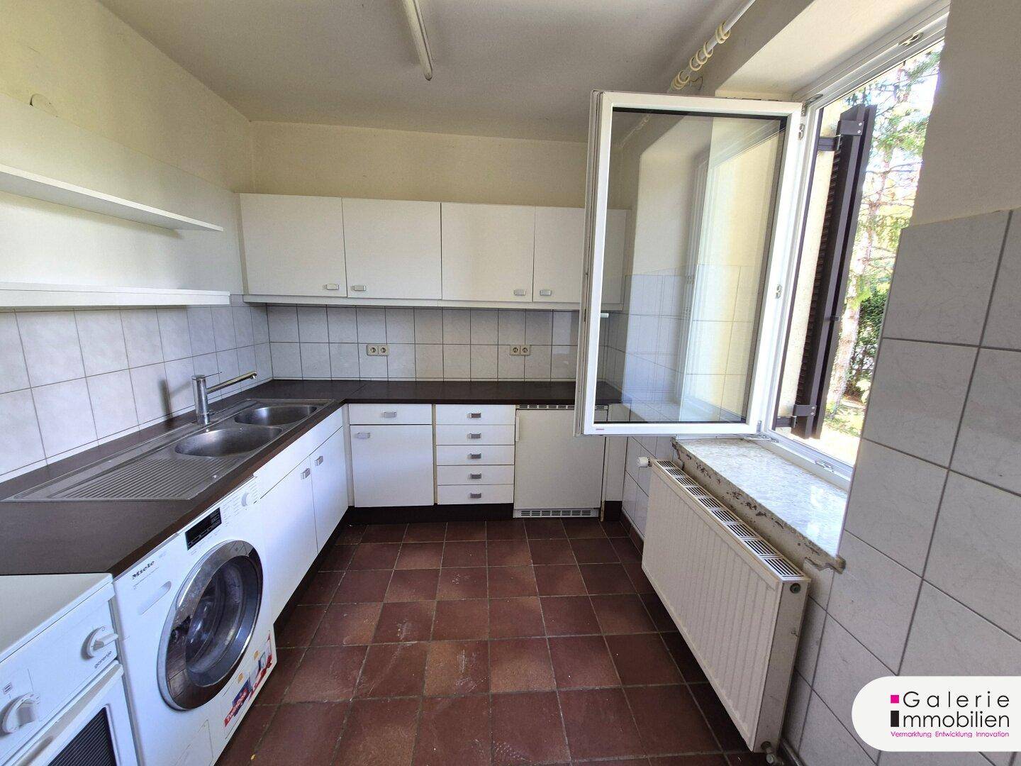 Einfache Küche mit weißen Schränken, Waschmaschine und Fenster mit Blick ins Grüne.