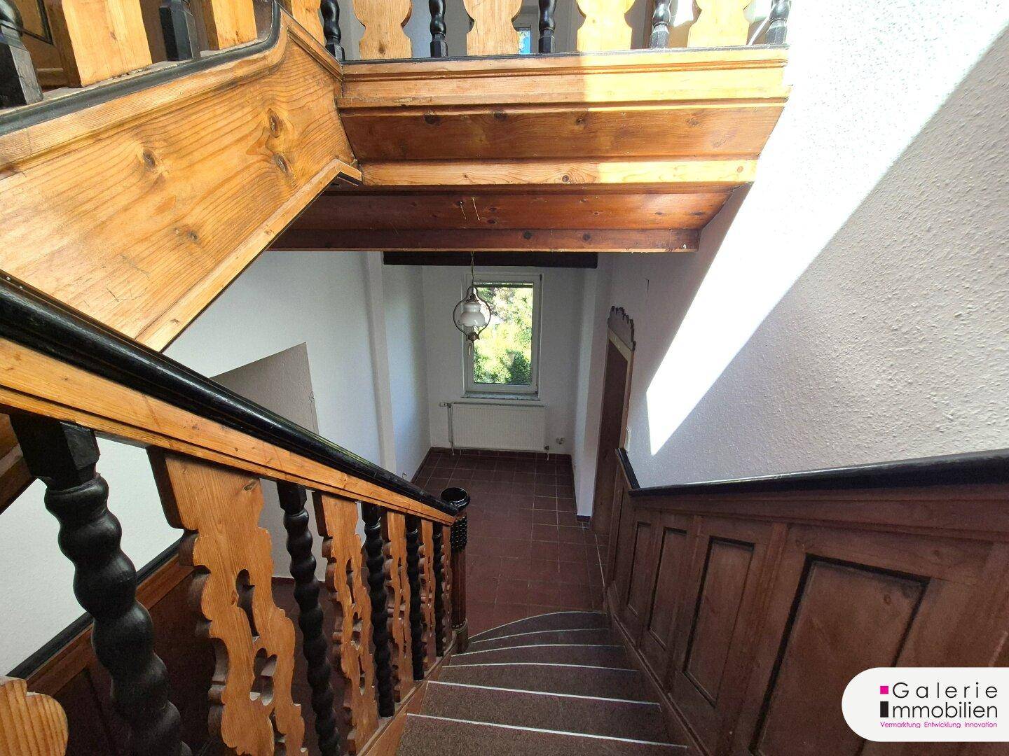 Traditionelles Holztreppenhaus mit geschnitzten Geländern und Blick auf den Garten durch das Fenster.