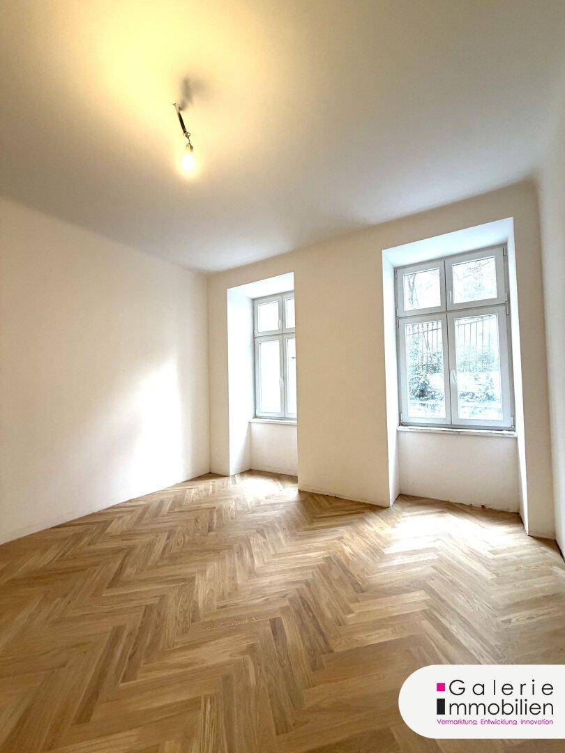 Helles Zimmer mit Fischgrätparkett und zwei großen Fenstern, bereit zur individuellen Gestaltung.