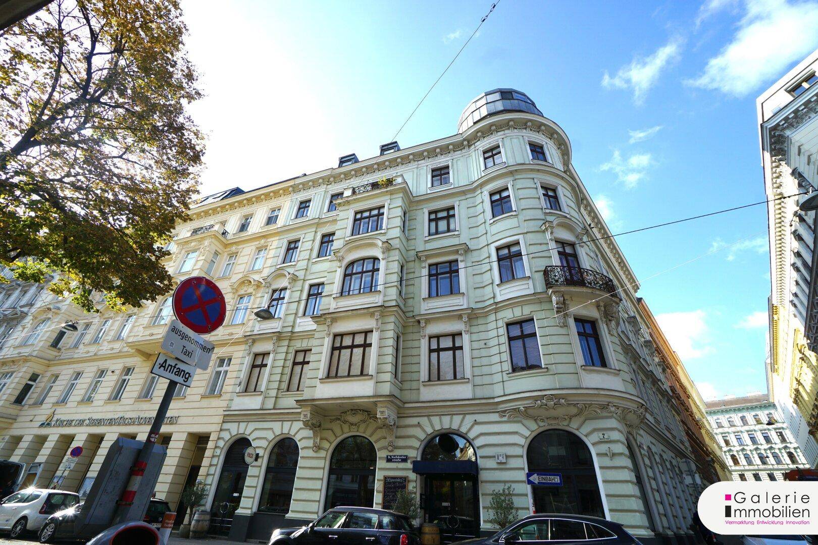 Prächtiges Eckgebäude mit historischer Fassade und großen Fenstern in einer sonnigen Stadtansicht.
