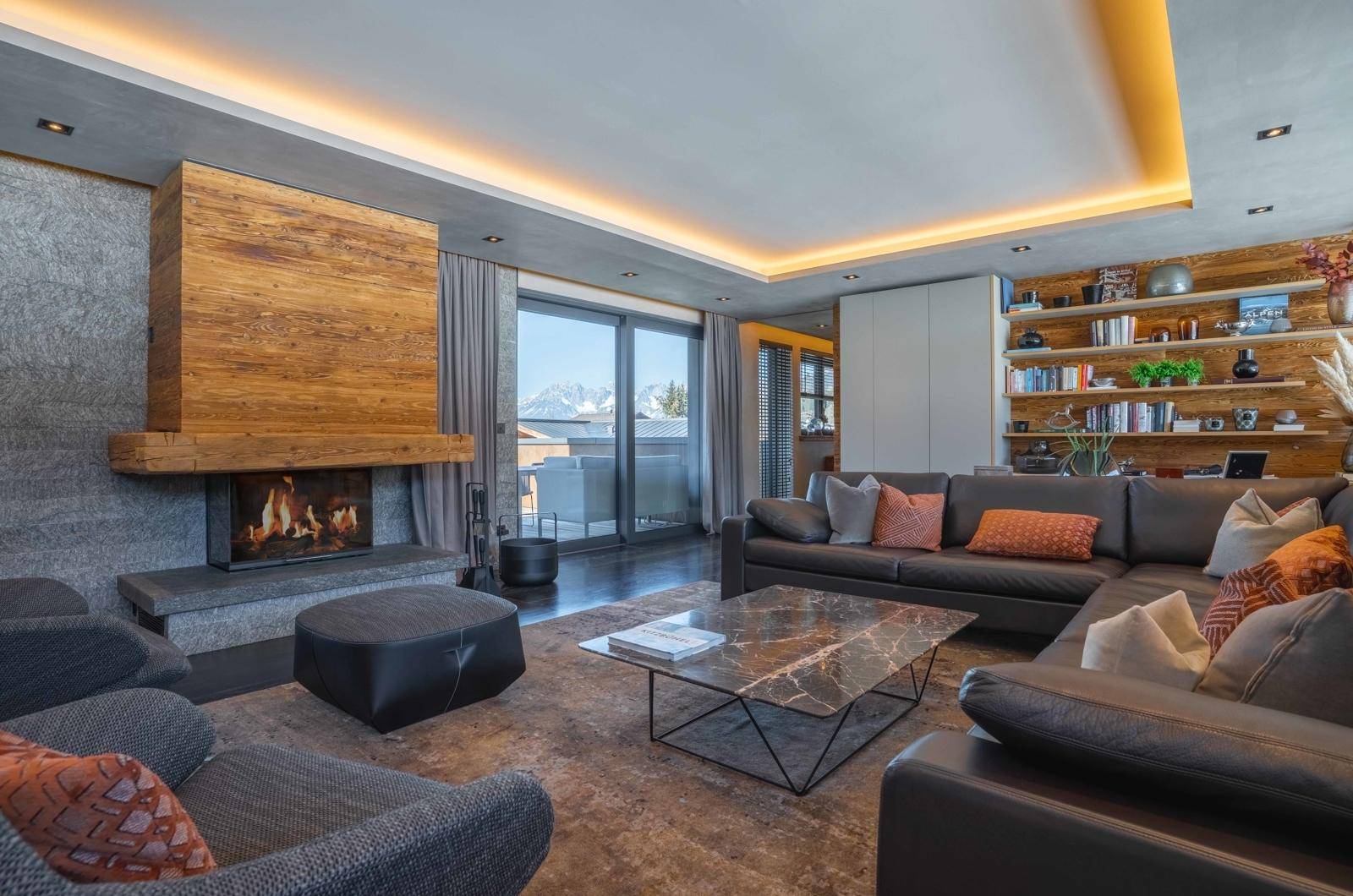 Elegantes Wohnzimmer mit Kamin, dunklem Sofa, Marmortisch und Zugang zum Balkon mit Bergblick.