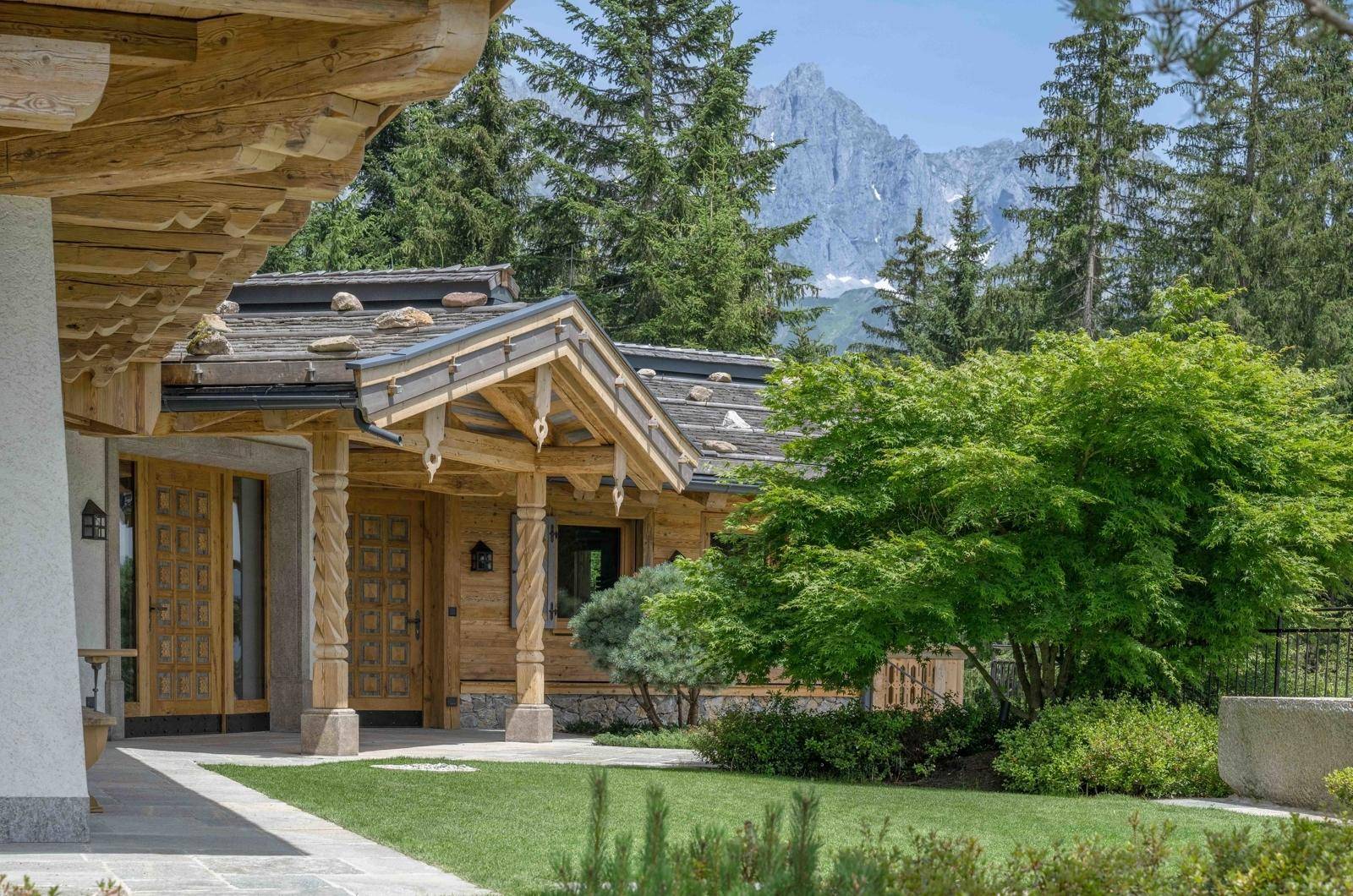 Eingangsbereich des Chalets mit kunstvoll geschnitzten Holzsäulen und gepflegtem Rasen vor Bergkulisse.