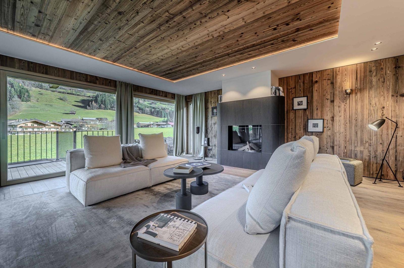 Helles Wohnzimmer mit Kamin, Holzwänden und Panoramablick auf die grüne Berglandschaft.