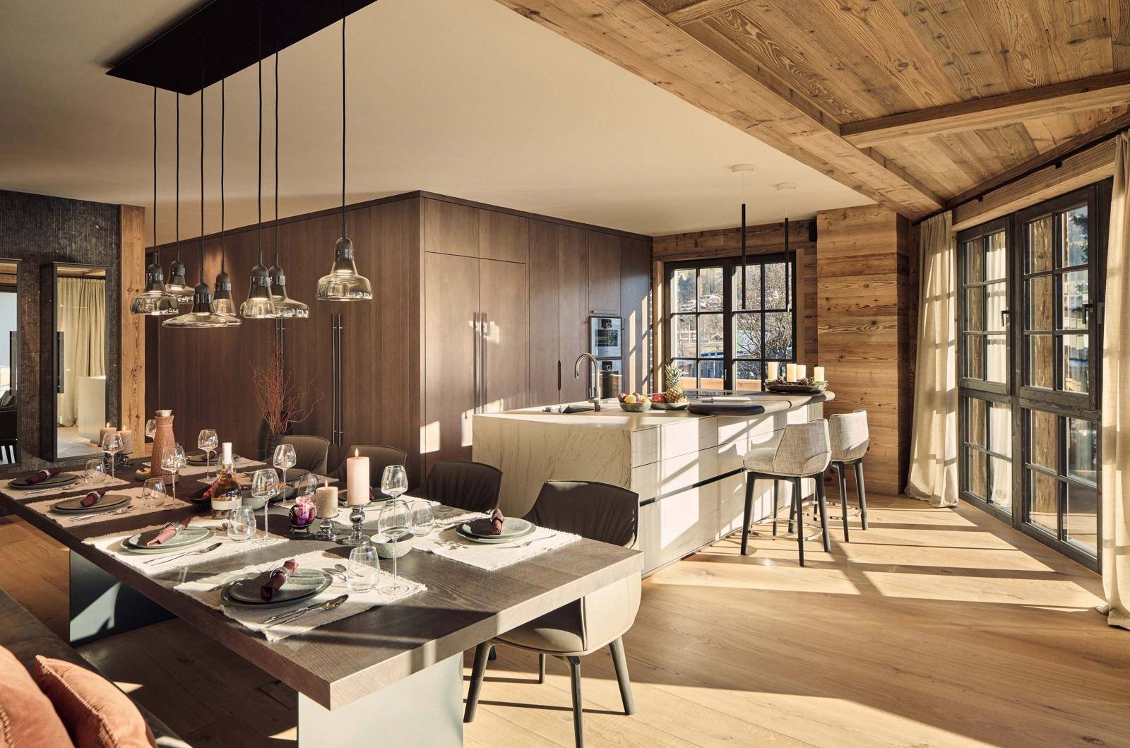 Offene Küche mit Essbereich, Holzschränken, Kücheninsel und großen Fenstern mit viel Tageslicht.