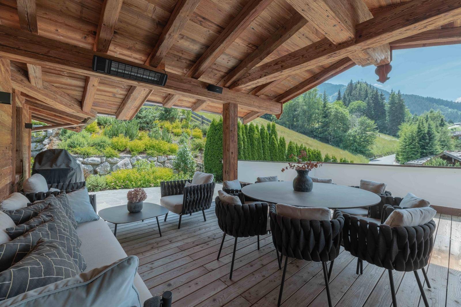Große überdachte Terrasse mit Essbereich und Lounge-Möbeln, umgeben von Natur und Bergen.