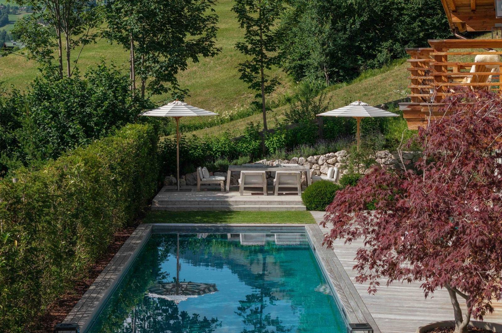 Idyllischer Poolbereich mit Holzterrasse, Sonnenschirmen und Essbereich im Grünen mit Bergblick.