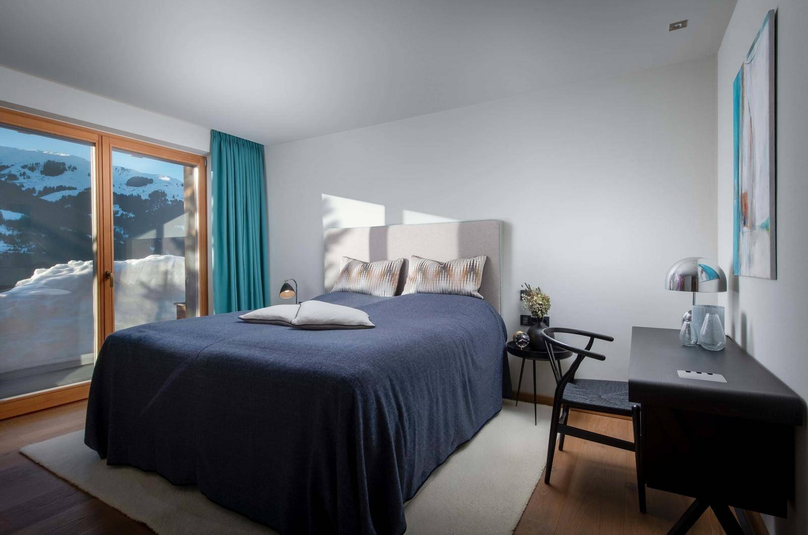 Komfortables Schlafzimmer mit Schreibtisch und Zugang zu einem Balkon mit Blick auf die verschneite Landschaft.