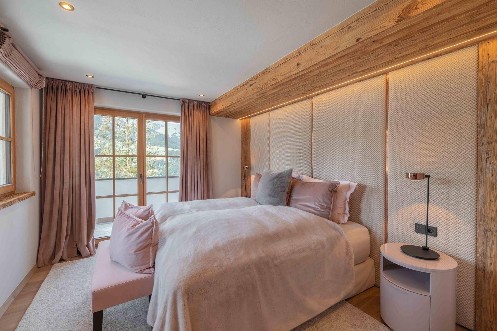 Gemütliches Schlafzimmer mit weichen Textilien, Holzbalken und Fensterblick in die Natur.