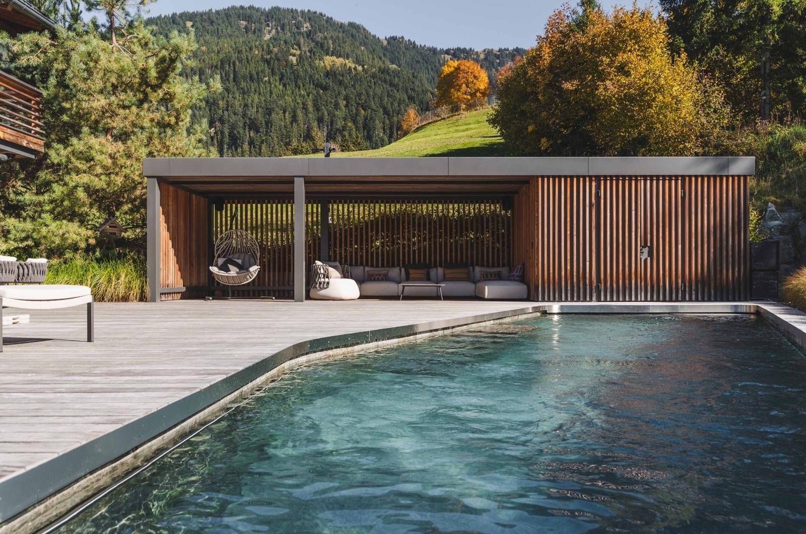 Moderner Außenpoolbereich mit Holzdeck, überdachter Lounge und bequemen Sitzgelegenheiten vor Bergkulisse.