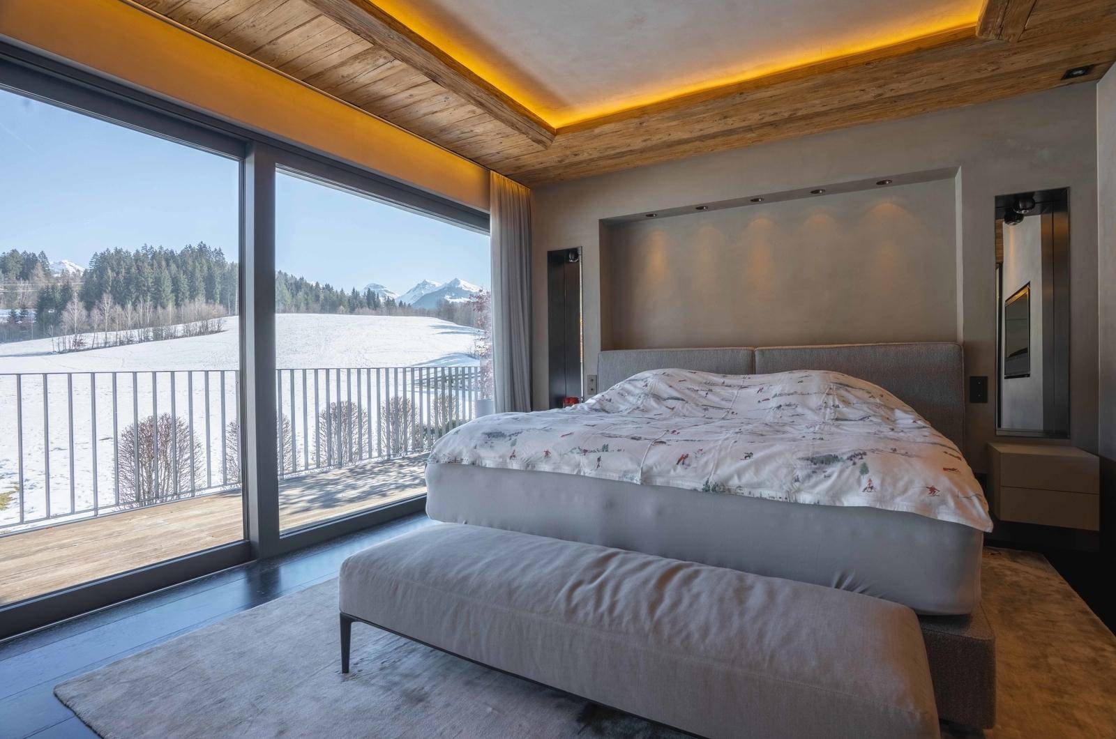 Helles Schlafzimmer mit großem Bett, Holzbalkendecke und bodentiefen Fenstern mit Winterlandschaftsblick.