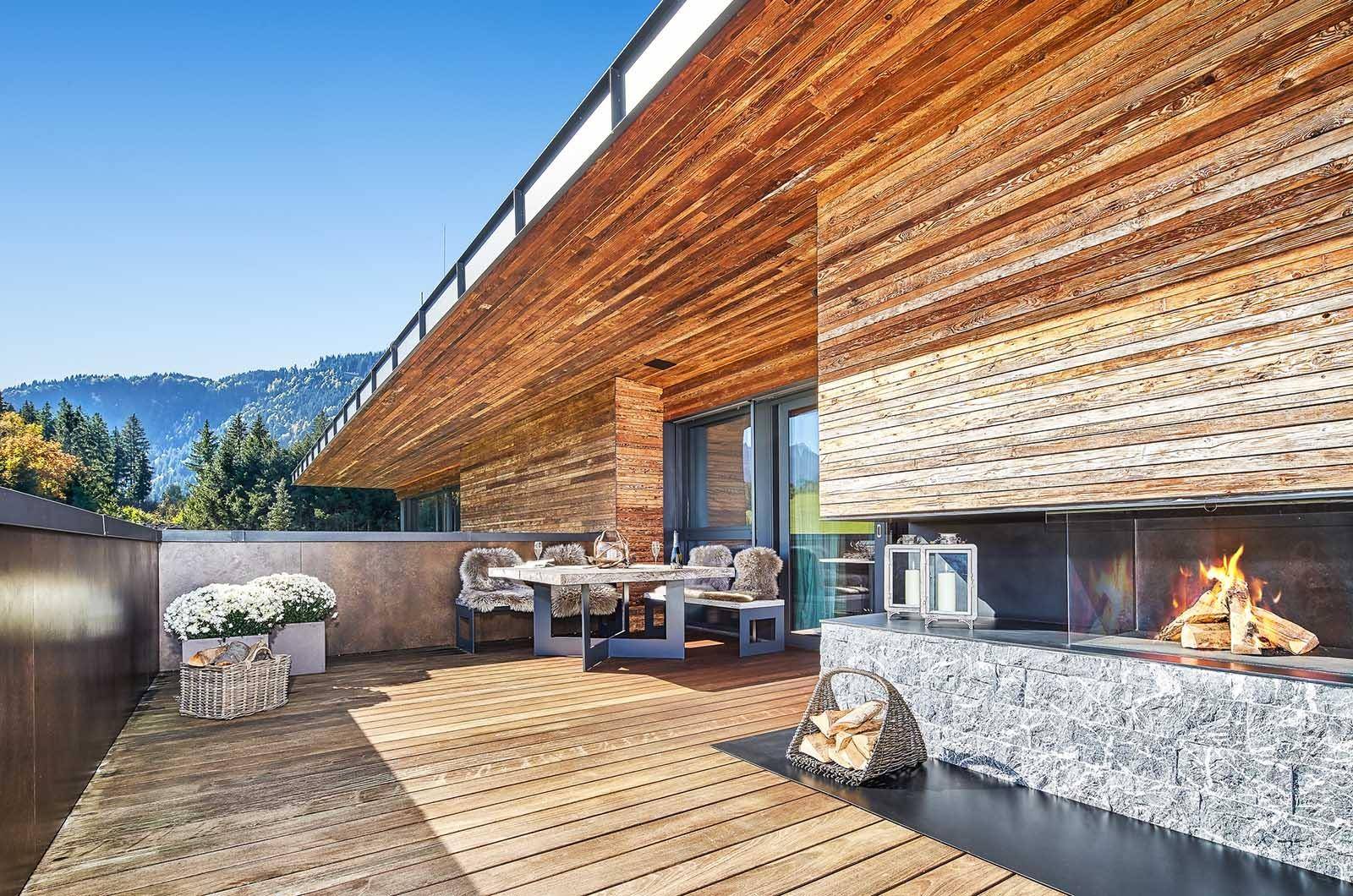 Sonnige Terrasse mit Holzboden, Esstisch und integriertem Außenkamin, umgeben von Natur.