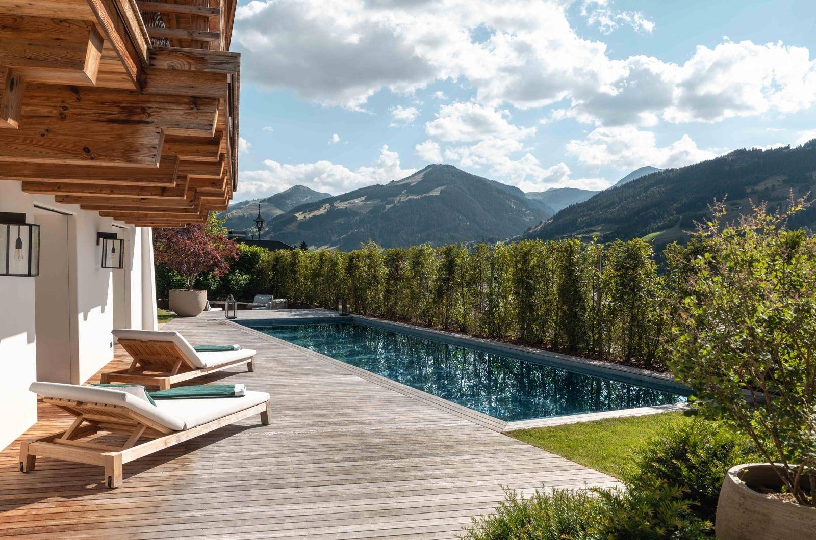 Entspannender Poolbereich mit Holzdeck, Sonnenliegen und atemberaubendem Bergblick unter blauem Himmel.