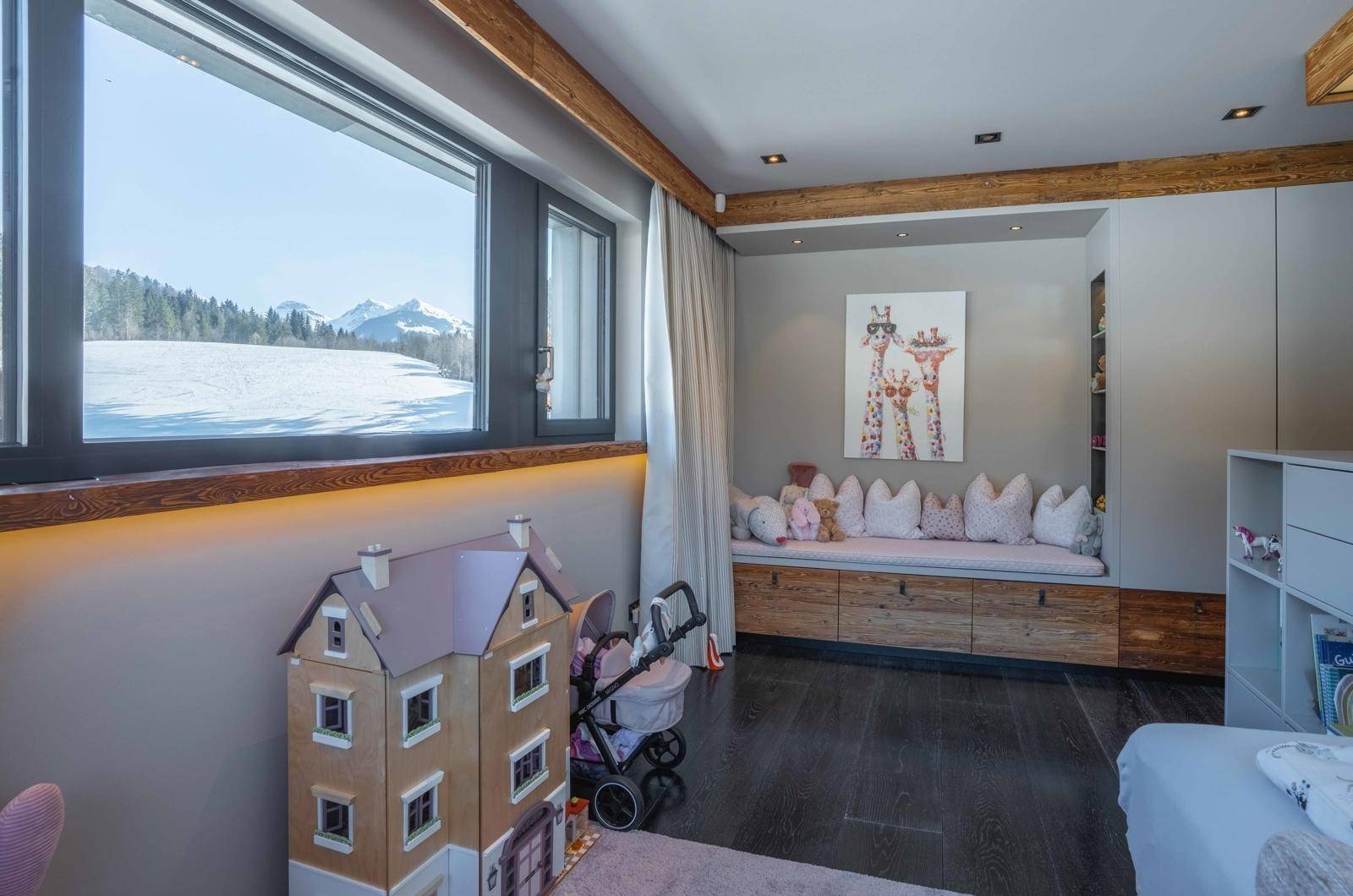 Kinderzimmer mit großem Fenster, Holzbalken und Spielzeug, bietet Blick auf die verschneite Landschaft.