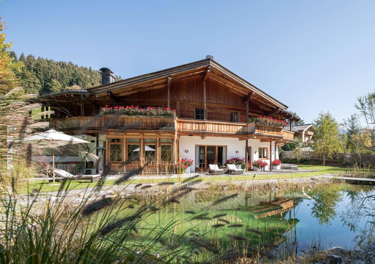 Traditionelles Holzchalet mit mehreren Balkonen, Terrasse und einem Naturteich im Vordergrund.