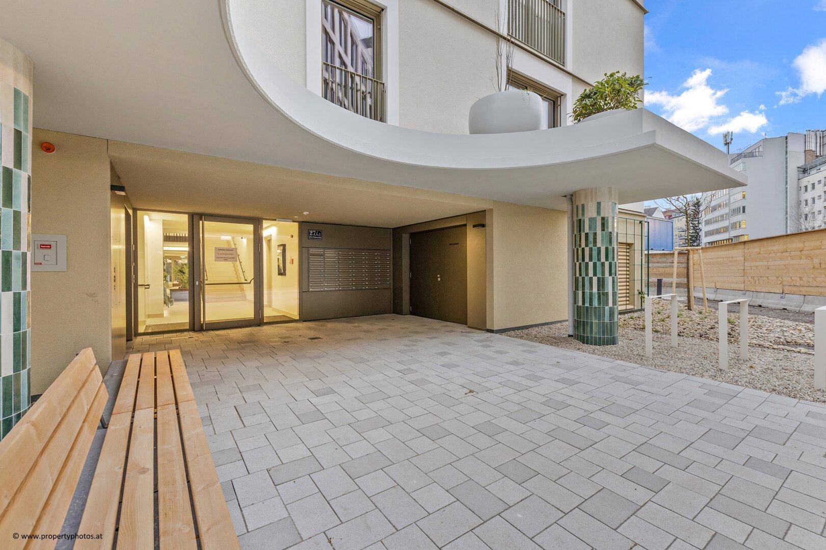 Eingangsbereich eines modernen Wohnhauses mit Briefkästen, gepflastertem Vorplatz und Sitzgelegenheit.