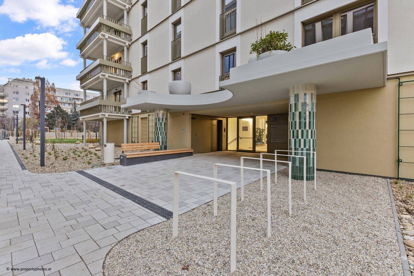 Außenansicht des Gebäudeeingangs mit Fahrradständern, Sitzbank und gepflegter Außenanlage.