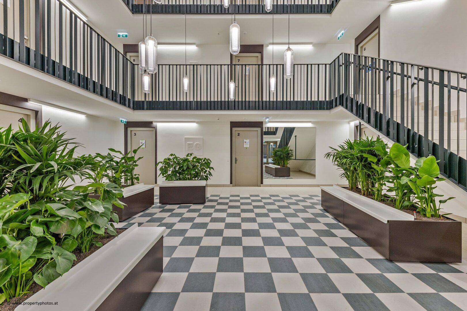 Heller, mehrstöckiger Eingangsbereich mit Schachbrettmuster-Boden, vielen Pflanzen und modernen Hängelampen.