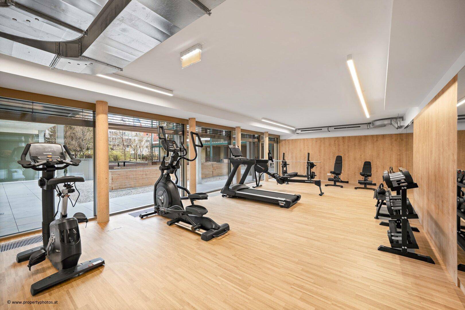 Gut ausgestatteter Fitnessraum mit verschiedenen Trainingsgeräten, Holzboden und großen Fenstern.