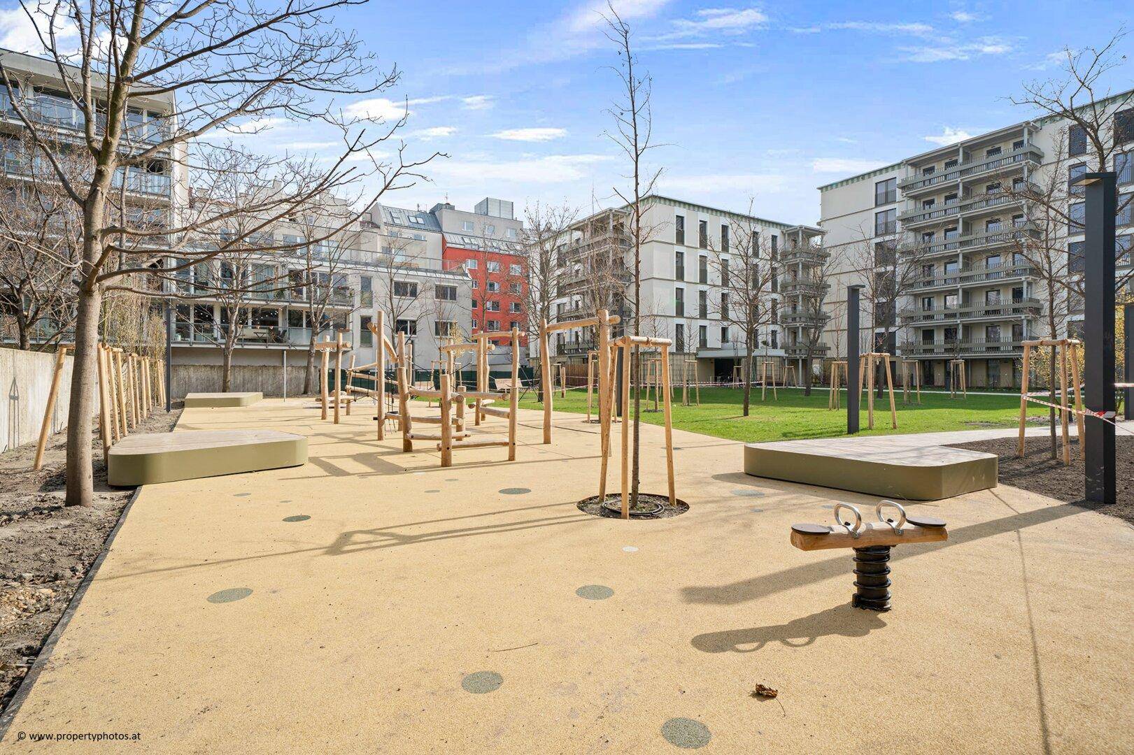 Großer Kinderspielplatz mit verschiedenen Holzspielgeräten und Sandfläche zwischen Wohngebäuden.