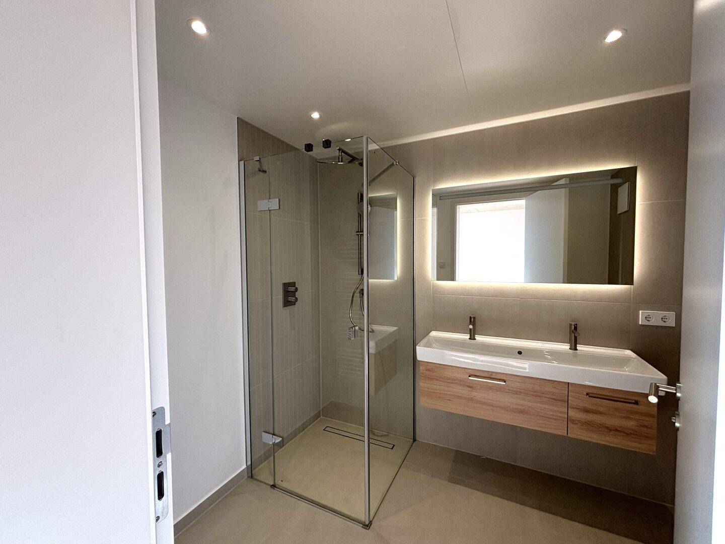Modernes Badezimmer mit begehbarer Dusche, Doppelwaschbecken und großem beleuchteten Spiegel.