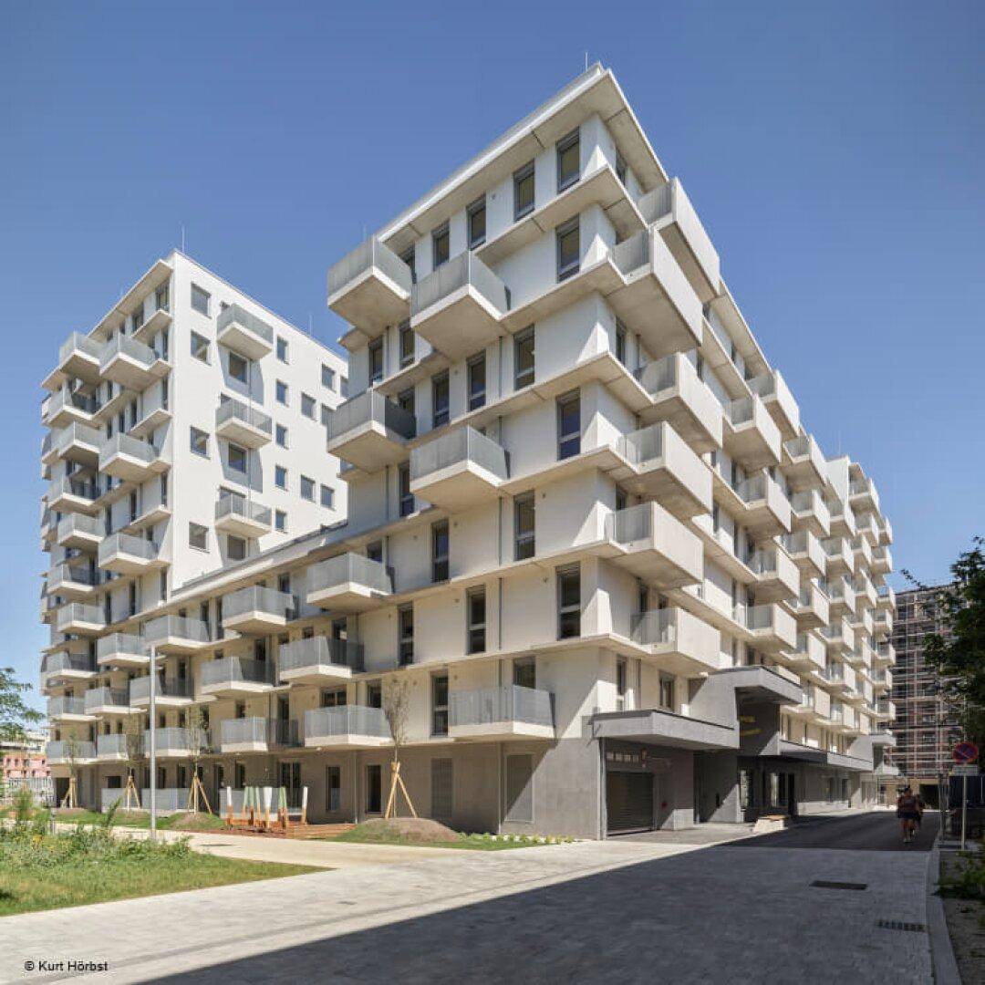 Zwei moderne Wohngebäude mit vielen Balkonen und klaren Linien unter strahlend blauem Himmel.