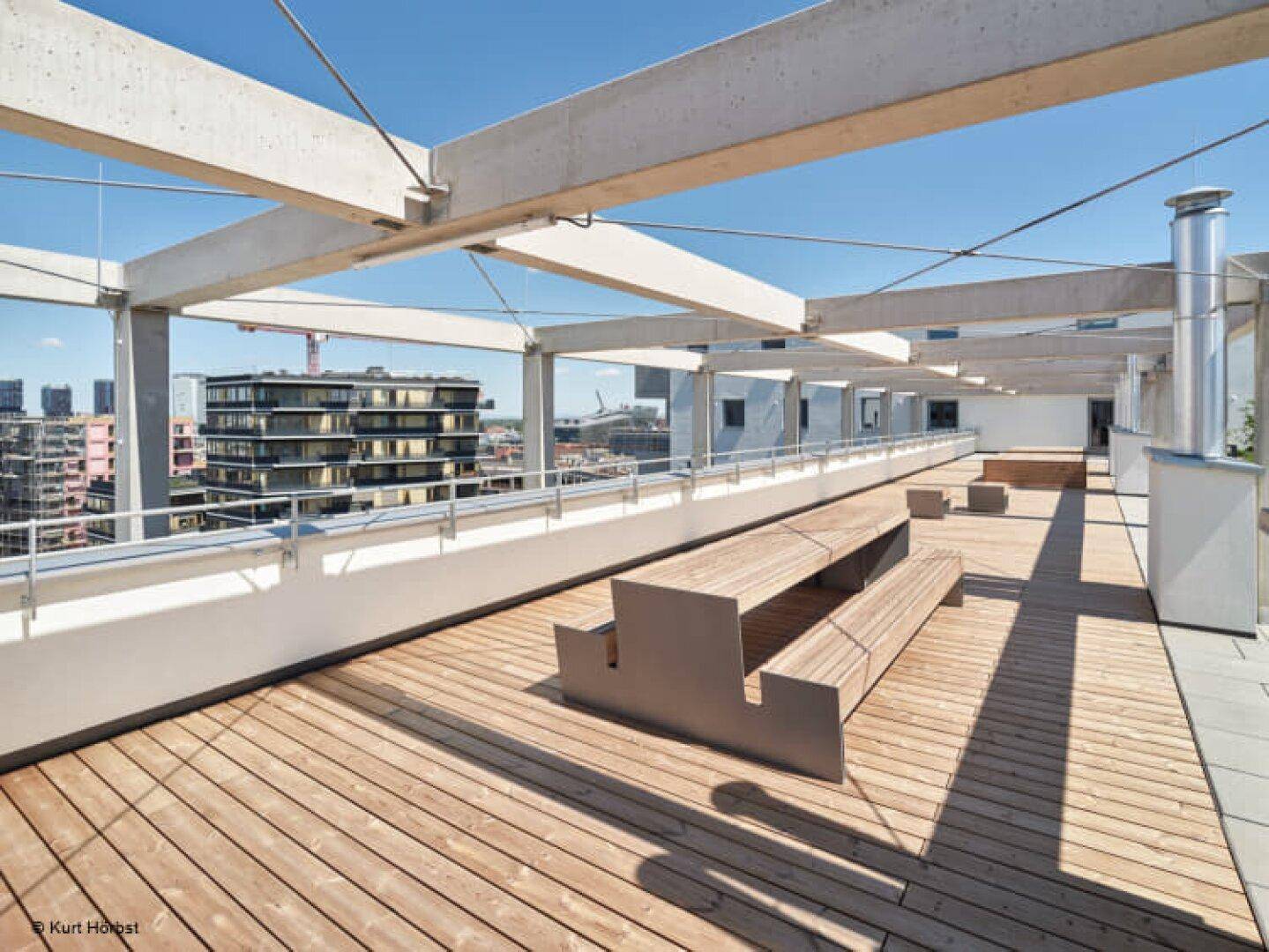 Weitläufige Dachterrasse mit Holzbänken und Tischen unter einer Betonpergola mit Stadtblick.