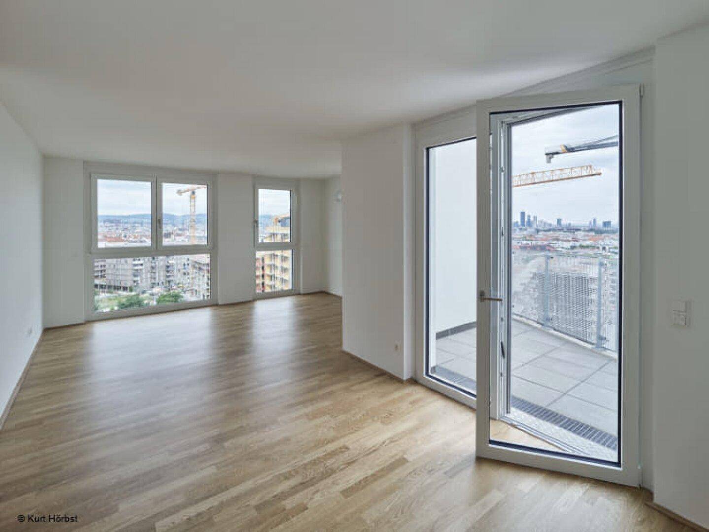 Geräumiges Wohnzimmer mit Parkettboden, großen Fenstern und Zugang zu einem Balkon mit Stadtblick.
