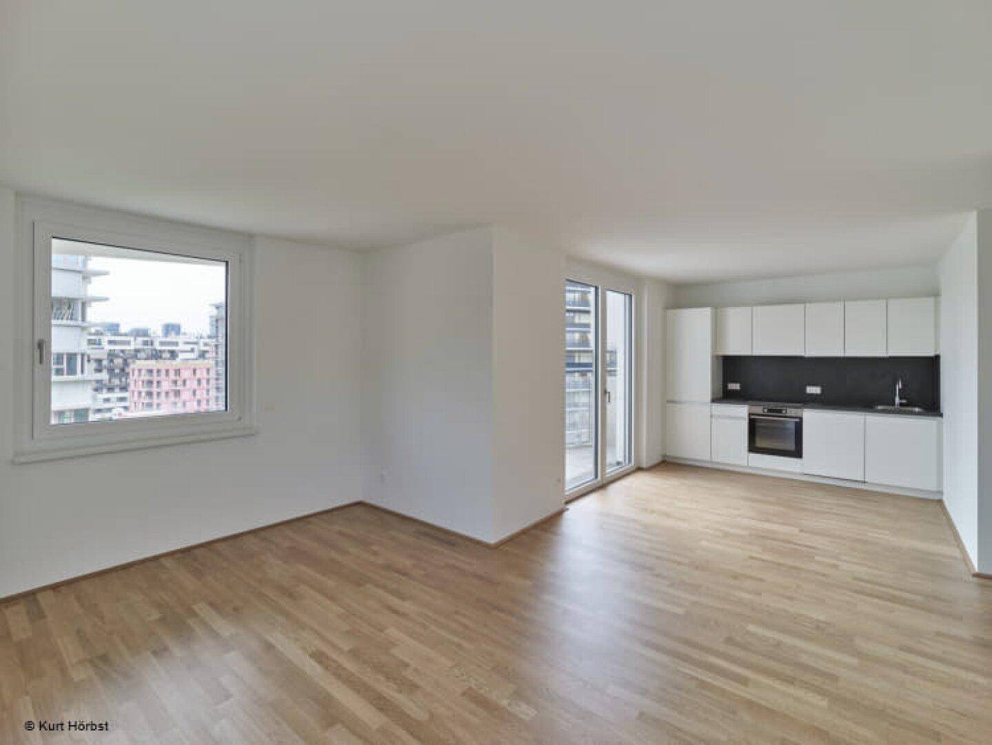 Offener Wohnbereich mit Parkettboden, weißer Küchenzeile und Zugang zu einem Balkon.