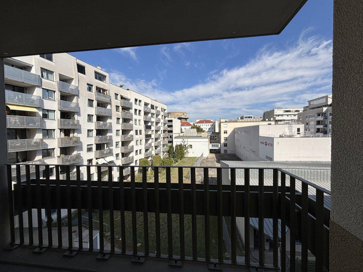 Blick vom Balkon auf einen begrünten Innenhof und umliegende moderne Wohngebäude unter blauem Himmel.