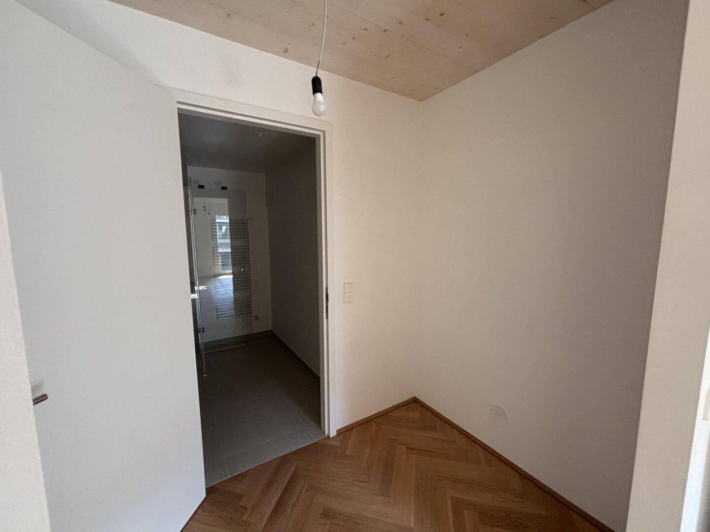 Helles Zimmer mit Fischgrätparkett und Blick in ein modernes Badezimmer, ideal für ein Schlafzimmer.