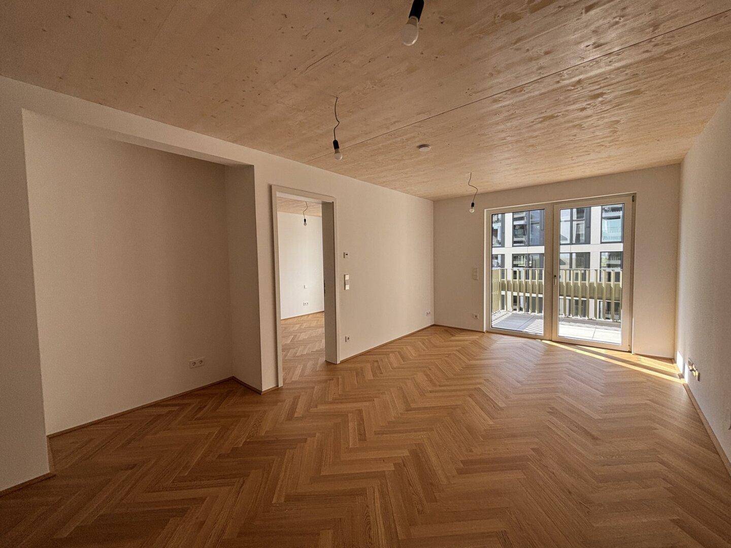 Geräumiger, heller Wohnbereich mit Fischgrätparkett, Holzakzenten an der Decke und Zugang zum Balkon.
