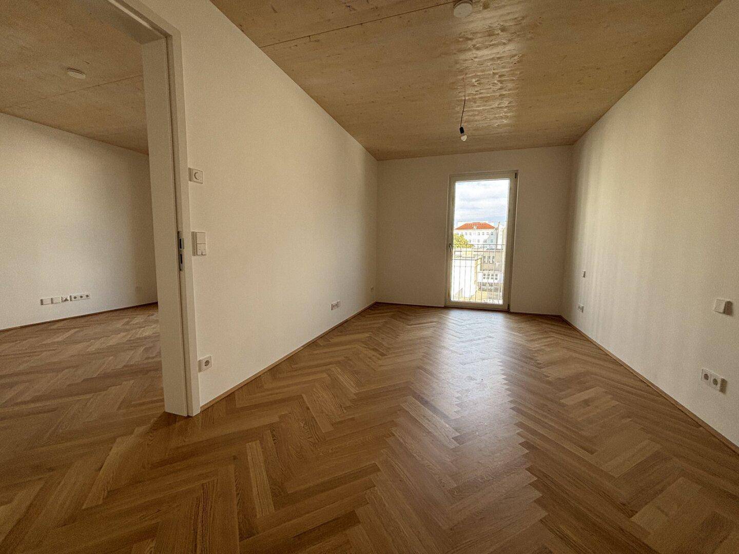 Helles, leeres Zimmer mit Fischgrätparkett, Holzbalkendecke und Zugang zu einem französischen Balkon.