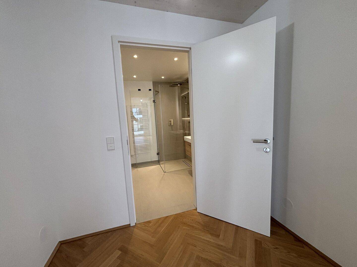 Heller Raum mit Fischgrätparkett und Blick in ein modernes Badezimmer mit Dusche.