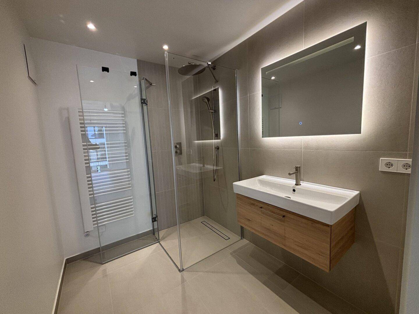 Modernes Badezimmer mit großer begehbarer Dusche, Waschtisch und beleuchtetem Spiegel.