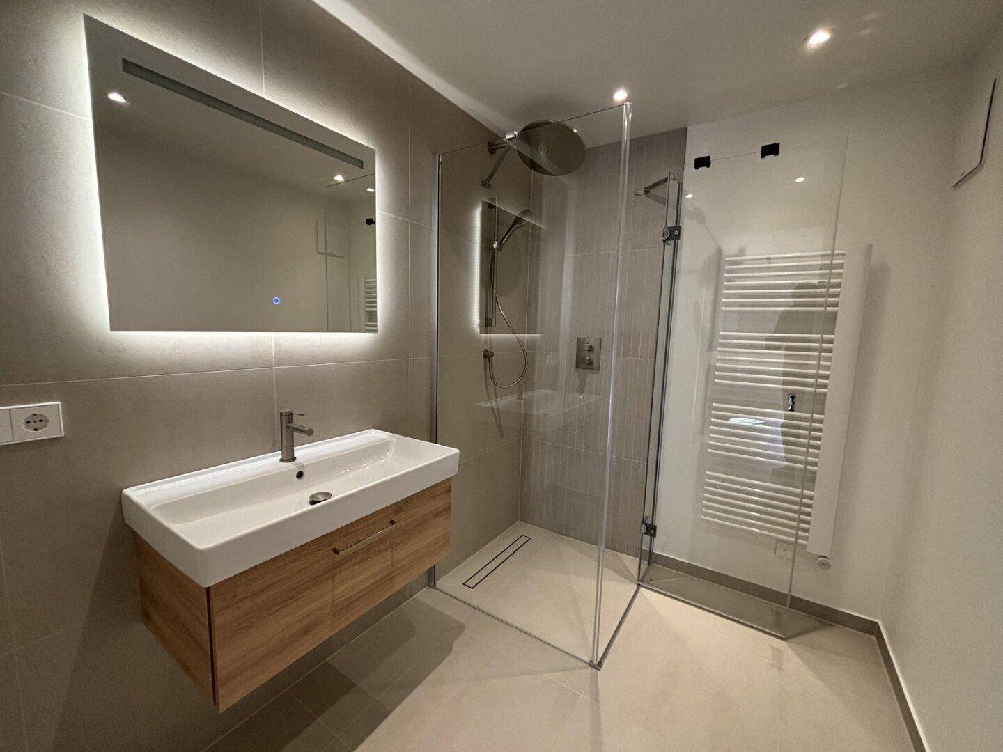 Modernes Badezimmer mit begehbarer Dusche, schwebendem Waschtisch und beleuchtetem Spiegel.