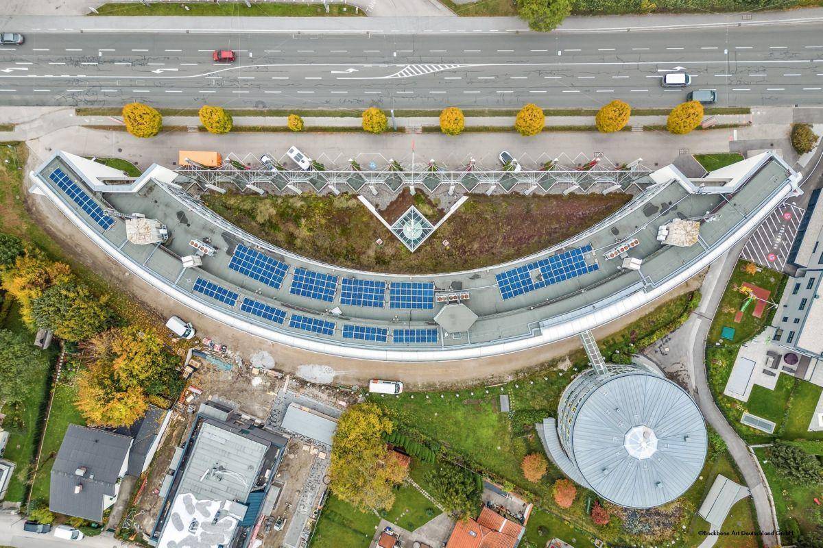 Senkrechte Luftaufnahme des geschwungenen Gebäudes mit Photovoltaikanlage auf dem Dach und begrünten Flächen.