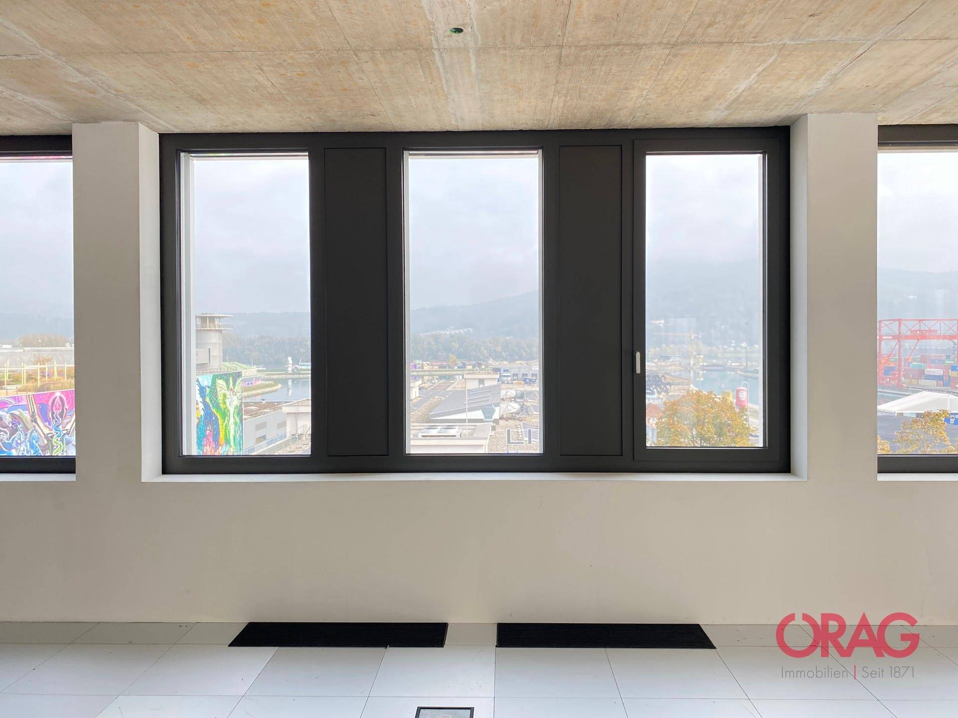Große Fensterfront mit Blick auf einen Hafenbereich und die städtische Umgebung, die viel Tageslicht hereinlässt.