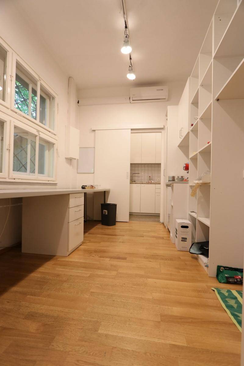 Helles Büro mit Holzfußboden, großen Fenstern, Einbauregalen und einer kleinen Küchenzeile im Hintergrund.