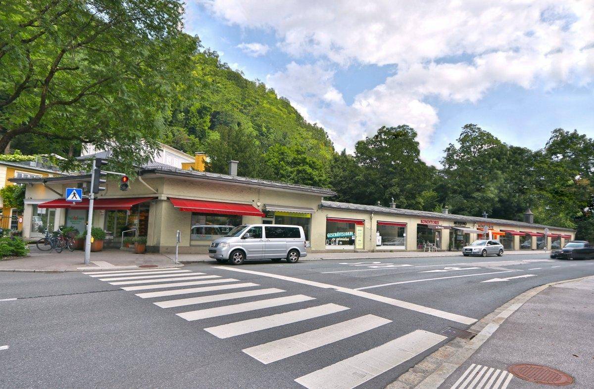 Belebte Geschäftsstraße mit mehreren Ladenlokalen, Fußgängerüberweg und grüner Hügellandschaft im Hintergrund.