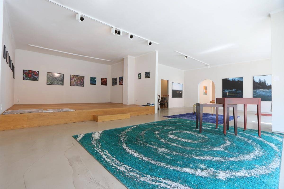 Geräumiger Ausstellungsbereich mit erhöhter Holzplattform, Kunstwerken und farbigen Teppichen auf dem Boden.