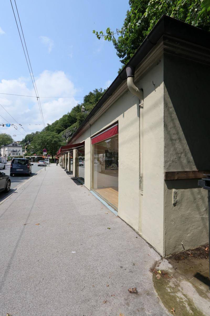 Seitenansicht eines Geschäftsgebäudes mit großen Schaufenstern und roter Markise entlang eines Bürgersteigs.