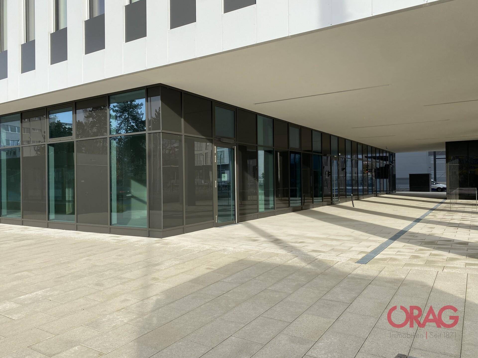 Moderne Gebäudefassade mit großen Glasflächen und überdachtem Eingangsbereich auf Straßenniveau.