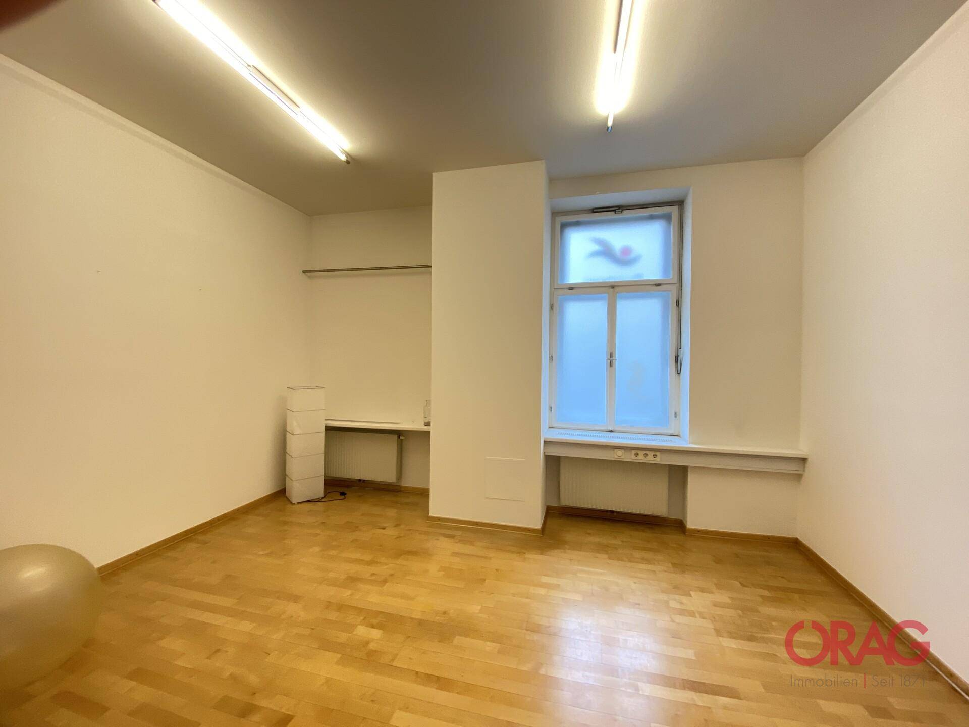 Geräumiger, heller Raum mit Holzparkett und großem Fenster, ideal für Büro oder Praxis.