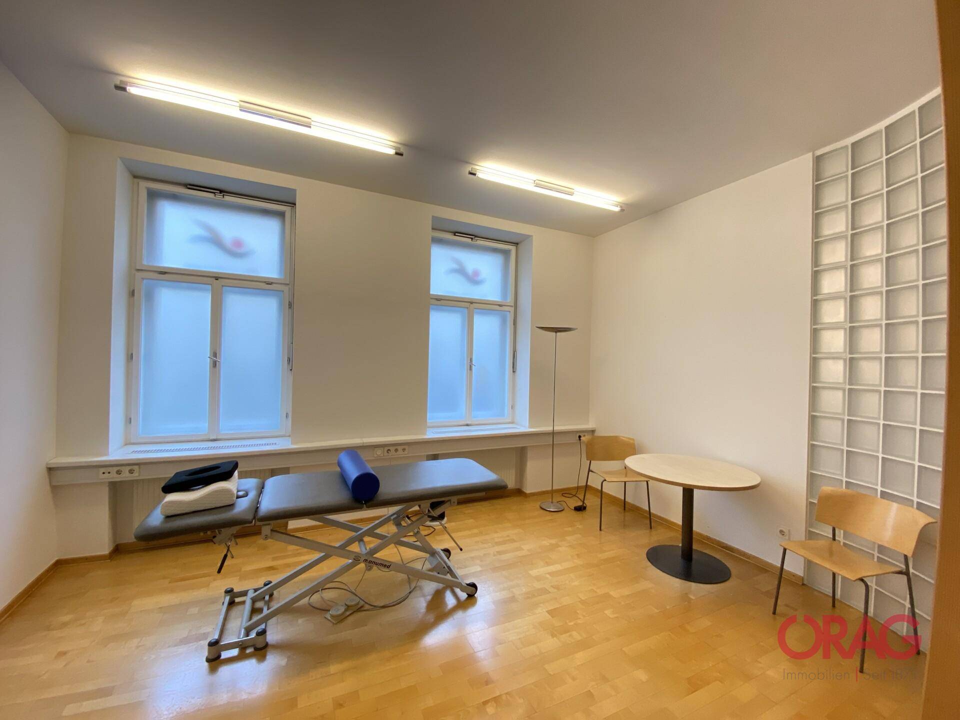 Praxisraum mit Behandlungsliege, zwei Fenstern und Holzfußboden, hell und funktional.