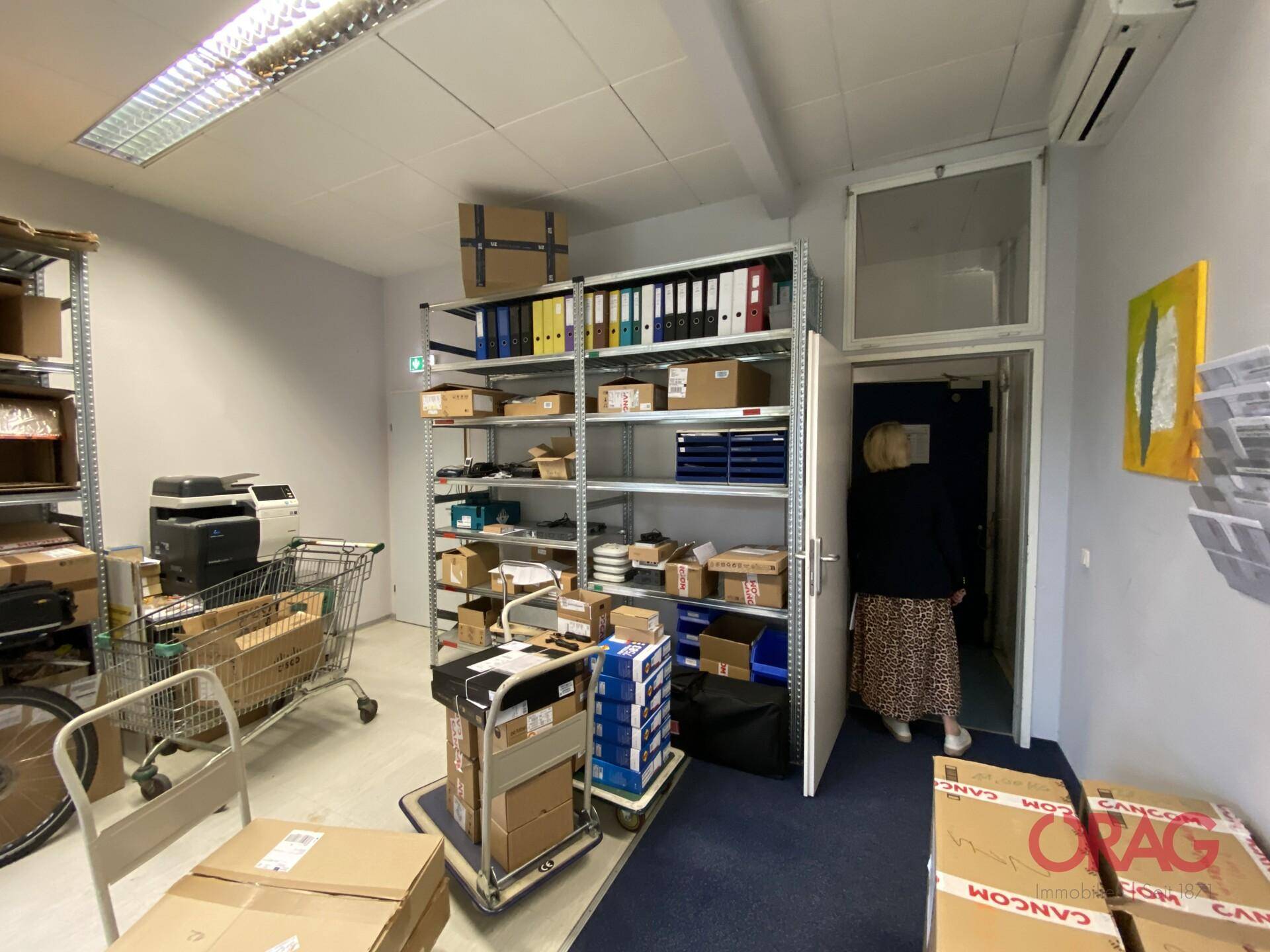 Büro-Lagerraum mit vollen Regalen, Kartons, einem Drucker und einem offenen Türbereich.