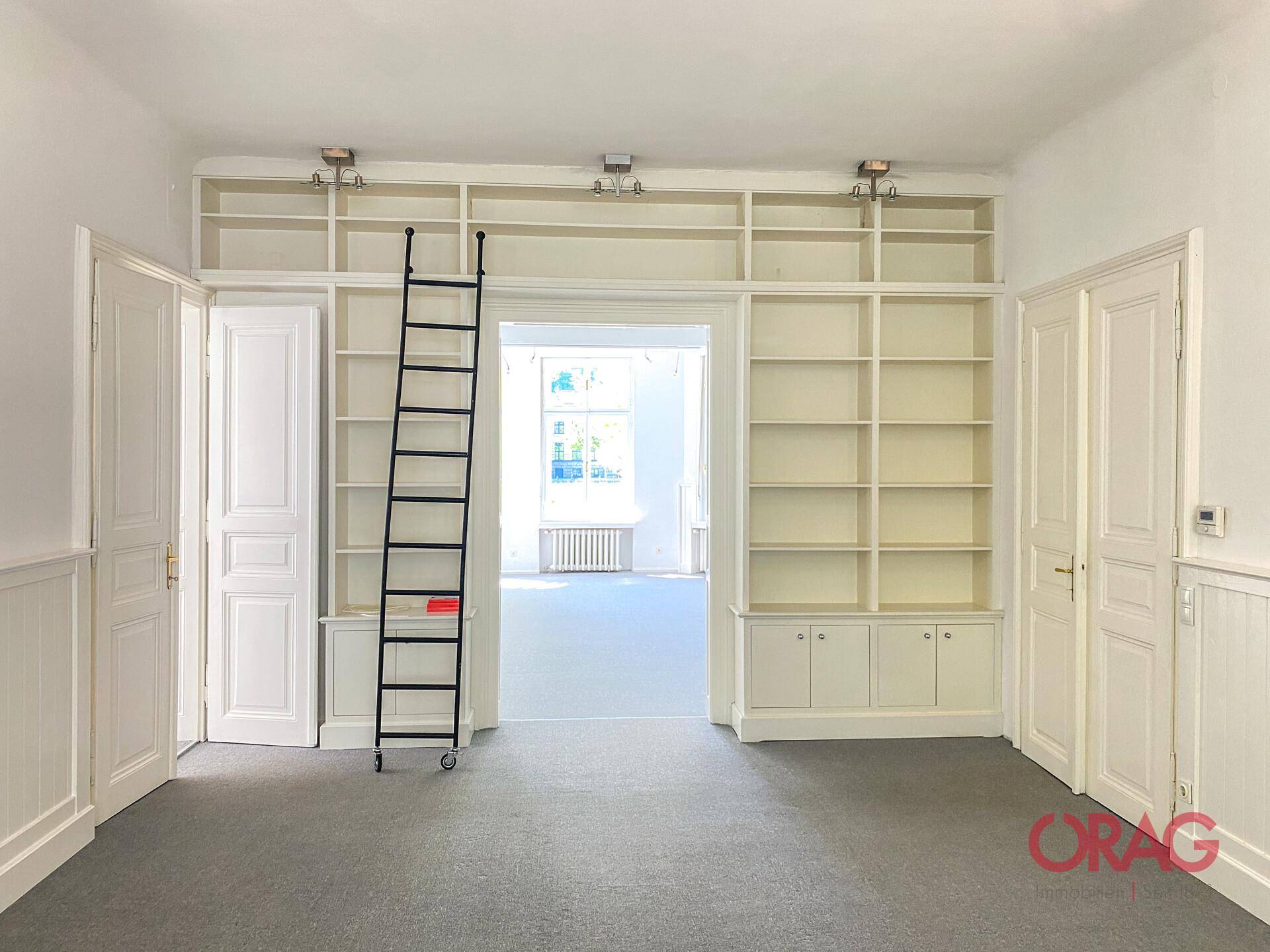 Geräumiger Raum mit eingebauten Bücherregalen, einer Leiter und Zugang zu einem weiteren Zimmer.